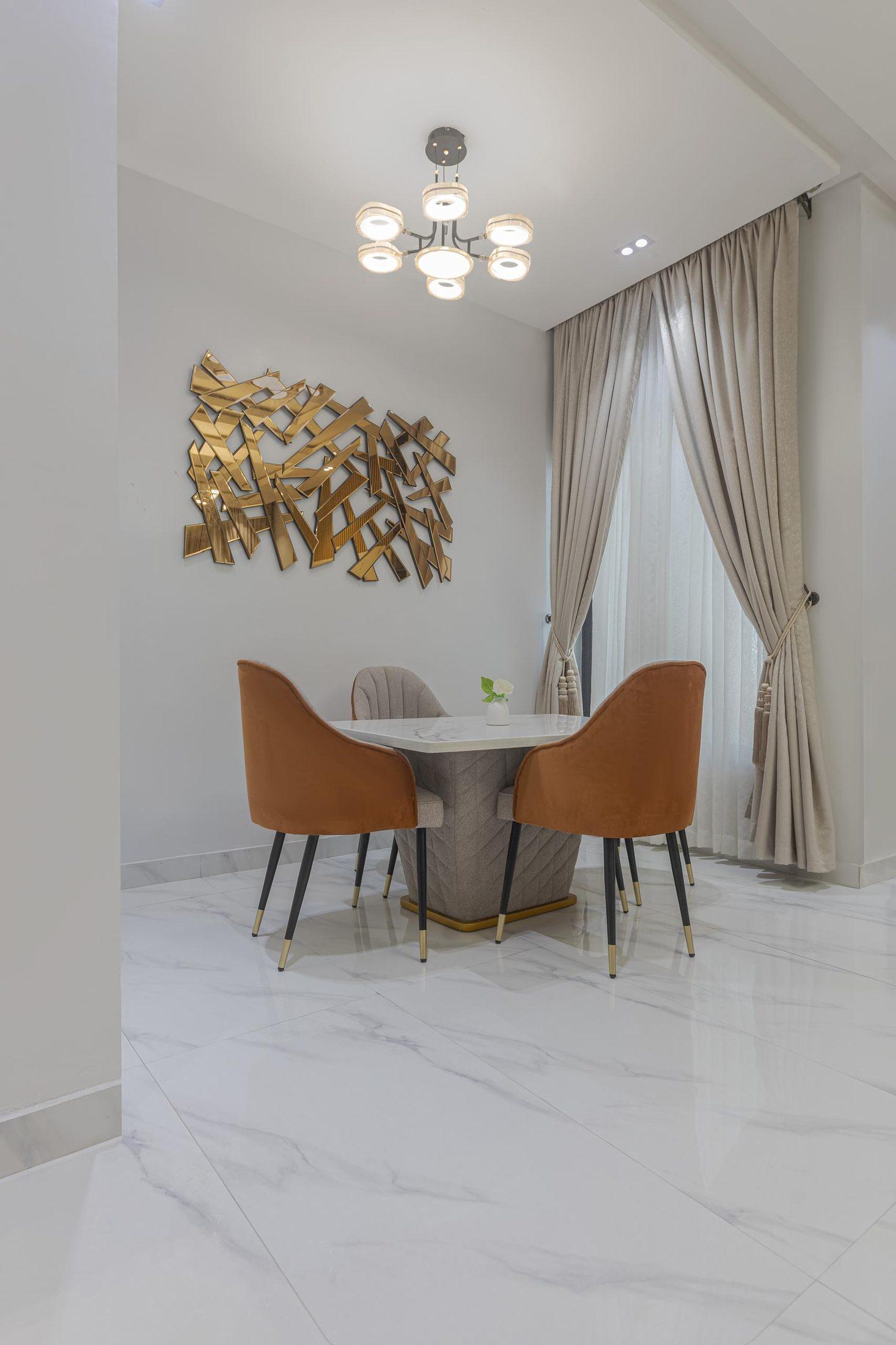 Round marble dining table with rust velvet chairs beneath a modern chandelier and a gold wall sculpture