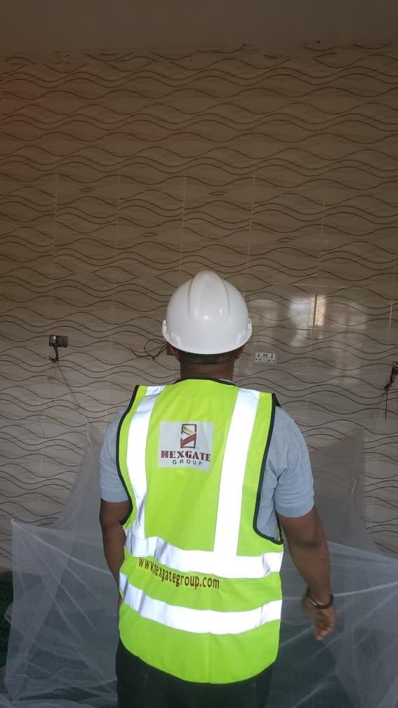 Nexgate team member seen from behind in a hi-vis vest printed with the Nexgate Group logo and nexgategroup.com, facing a tiled feature wall with sockets fitted