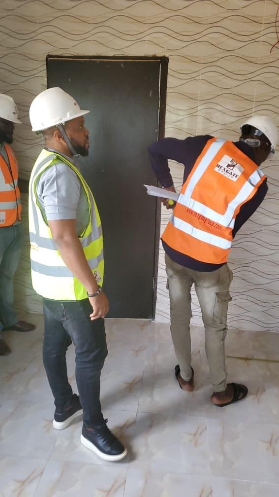 A Nexgate inspector bends to examine a door frame against a tiled wall while a colleague in a hi-vis vest watches
