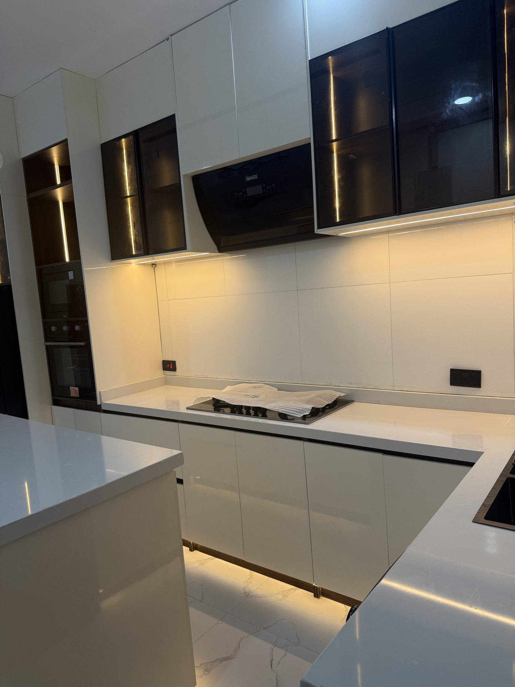 Hob wall with lit smoked glass wall units, a black cooker hood and built-in ovens in a cream gloss kitchen